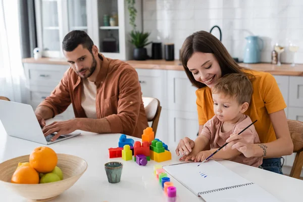 Mãe e filho desenhando perto de blocos de construção e marido usando laptop em casa — Fotografia de Stock