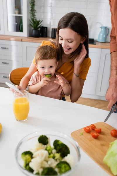 Mãe feliz segurando smartphone e olhando para a criança comendo brócolis em casa — Fotografia de Stock