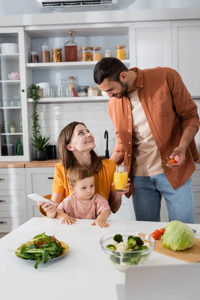 Uomo che tiene pomodori ciliegini vicino alla famiglia con smartphone e succo d'arancia a casa — Foto stock