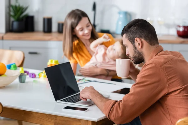 Hombre sosteniendo la taza y el uso de portátil cerca de la esposa y el hijo en casa - foto de stock