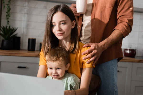 Man with cup hugging wife using laptop near son at home — Stock Photo