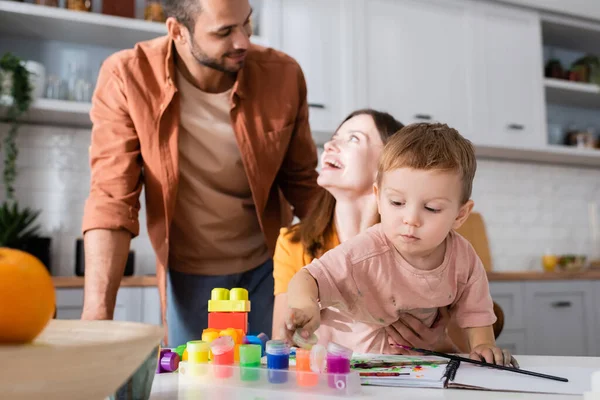 Enfant tout-petit tenant de la peinture près des blocs de construction et des parents à la maison — Photo de stock