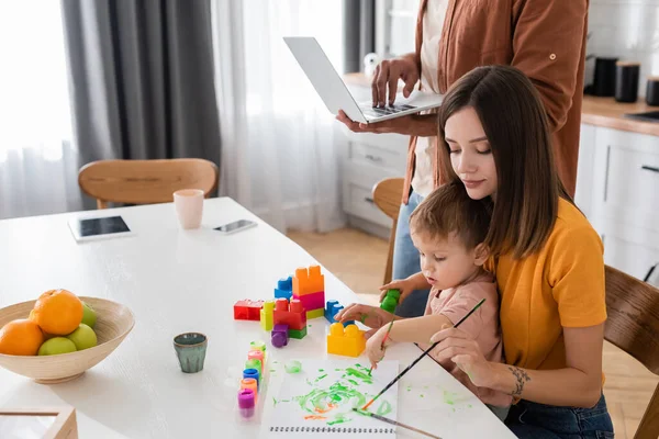 Frau zeichnet mit Sohn in der Nähe von Bauklötzen und Ehemann benutzt Laptop zu Hause — Stockfoto