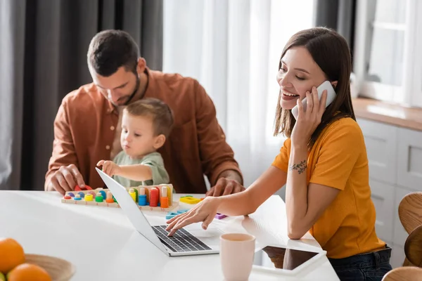 Femme souriante parlant sur smartphone et utilisant un ordinateur portable près du mari et de l'enfant à la maison — Photo de stock