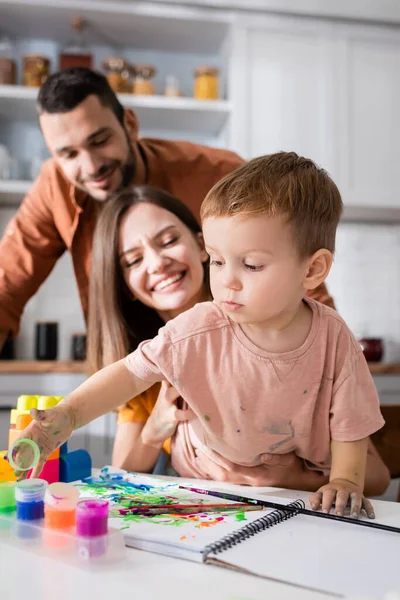 Niño pequeño jugando con pintura cerca de cuaderno de bocetos y los padres borrosos en casa - foto de stock