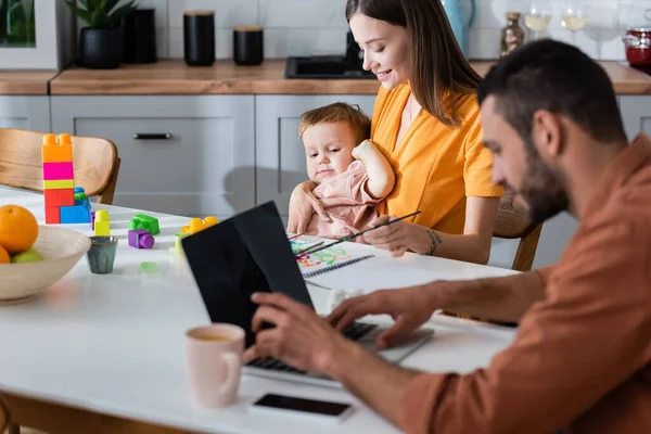 Mujer abrazando niño pequeño cerca de bloques de construcción y marido usando el ordenador portátil en casa - foto de stock