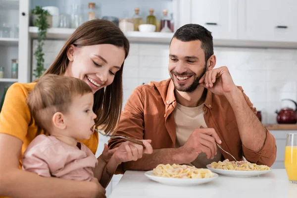 Sonriente hombre mirando esposa alimentación hijo con pasta cerca de jugo de naranja en cocina - foto de stock