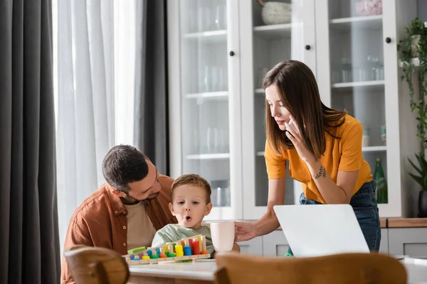 Mujer hablando en el teléfono inteligente cerca de la computadora portátil, marido e hijo jugando juego educativo - foto de stock