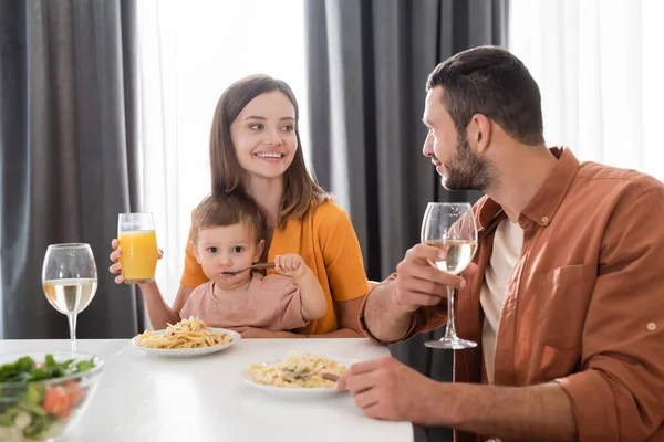 Mujer sonriente sosteniendo jugo de naranja cerca de hijo comiendo pasta y marido en casa - foto de stock