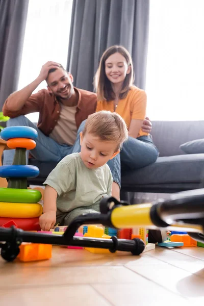 Малыш сидит рядом с игрушками и велосипедом рядом с родителями в гостиной — стоковое фото