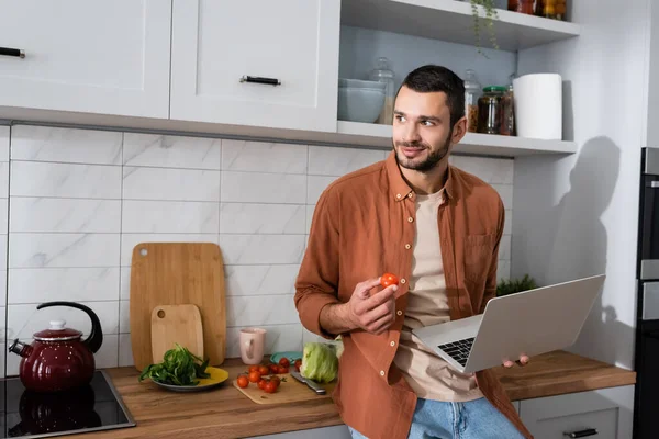 Smiling man holding laptop and cherry tomato in kitchen — Stock Photo