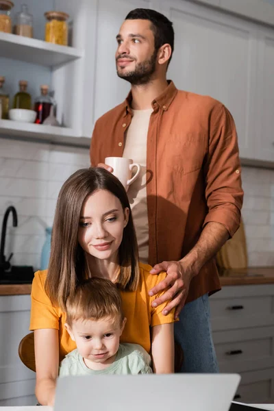 Giovane donna e bambino utilizzando computer portatile vicino all'uomo con tazza a casa — Foto stock