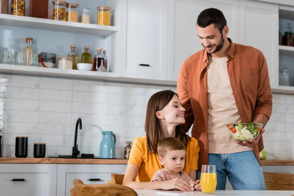 Uomo sorridente che tiene l'insalata vicino alla famiglia e il succo d'arancia in cucina — Foto stock