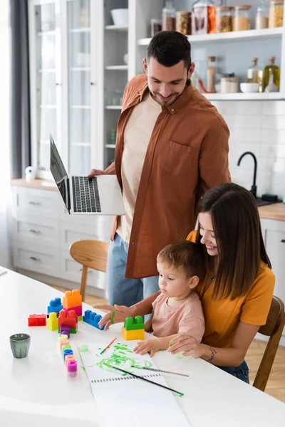 Uomo che tiene il computer portatile vicino alla famiglia giocando mattoni a casa — Foto stock