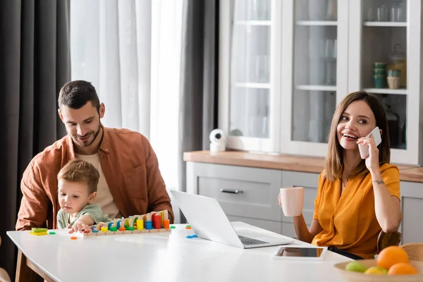 Freelancer sonriente hablando en smartphone cerca de la familia en casa - foto de stock