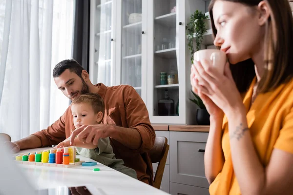 Man and kid playing educational game near woman with cup and digital tablet at home — Stock Photo