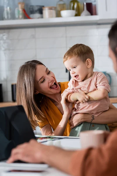 Excited mom hugging son near blurred husband using laptop at home — Stock Photo