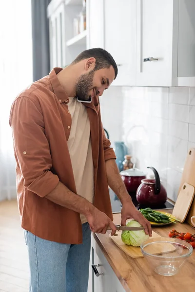 Uomo sorridente che parla sul telefono cellulare mentre taglia cavolo in cucina — Foto stock