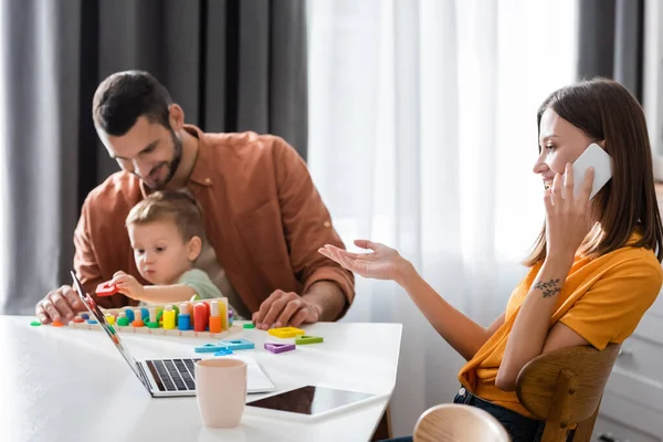 Smiling woman talking on cellphone near gadgets and blurred family at home — Stock Photo