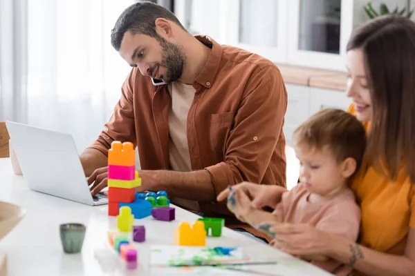 Uomo utilizzando il computer portatile e parlando su smartphone vicino alla famiglia con blocchi di costruzione a casa — Foto stock