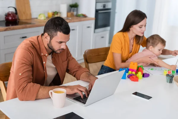 Young freelancer using laptop near coffee and family playing at home — Stock Photo