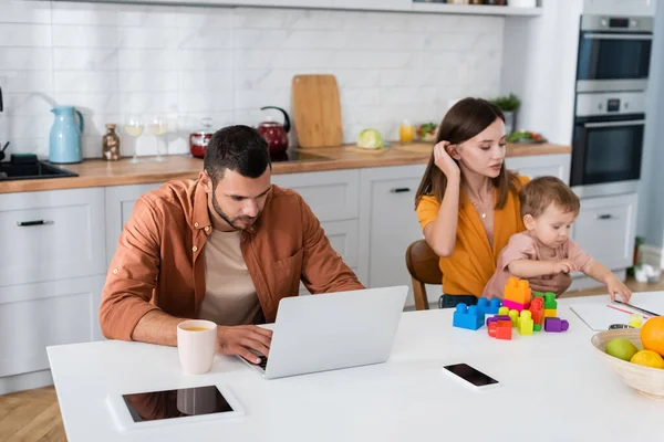 Hombre usando el ordenador portátil cerca de gadgets y esposa con hijo jugando bloques de construcción en casa - foto de stock