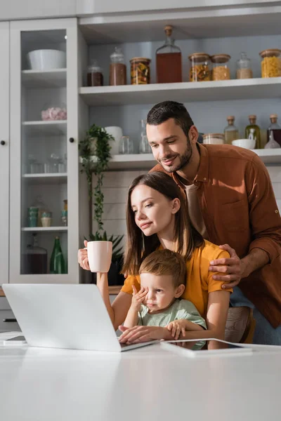 Hombre abrazando esposa con taza cerca de gadgets e hijo en casa - foto de stock