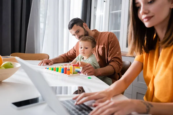 Hombre y niño jugando juego educativo cerca de la madre usando el ordenador portátil en casa - foto de stock