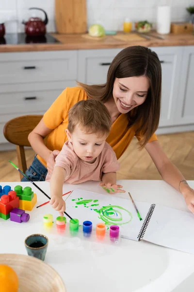 Femme souriante assise près du fils dessinant à la maison — Photo de stock