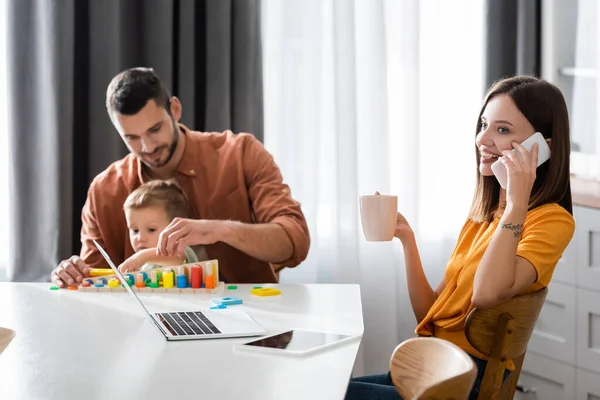 Smiling woman talking on cellphone and holding cup near laptop and family at home — Stock Photo