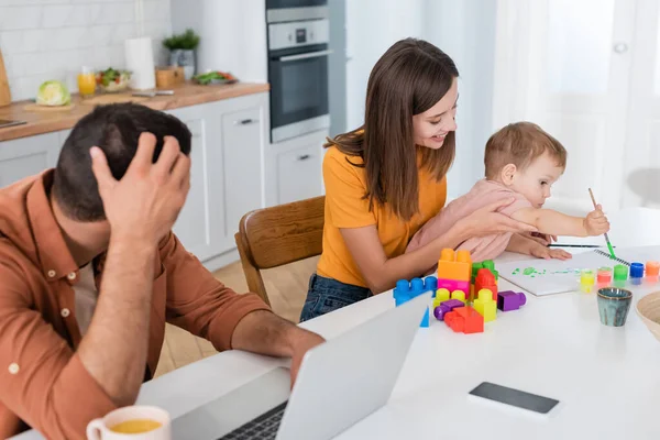 Enfant jouant près de sourire maman et père en utilisant un ordinateur portable à la maison — Photo de stock