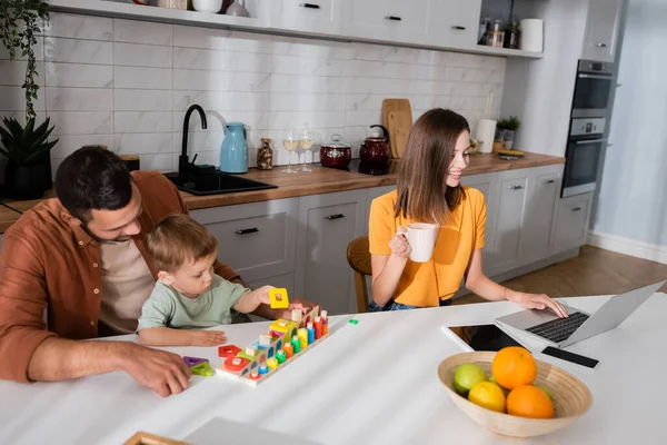 Freelancer con taza usando laptop mientras marido e hijo juegan juego en la cocina - foto de stock