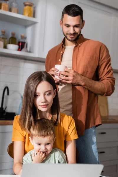 Junge Freiberuflerin schaut auf Laptop neben Sohn und verschwommenem Ehemann mit Tasse zu Hause — Stockfoto