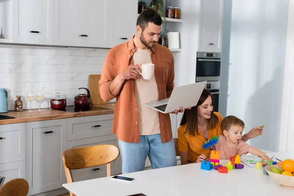 Mujer positiva jugando con el hijo cerca del marido con la taza y el ordenador portátil en casa - foto de stock