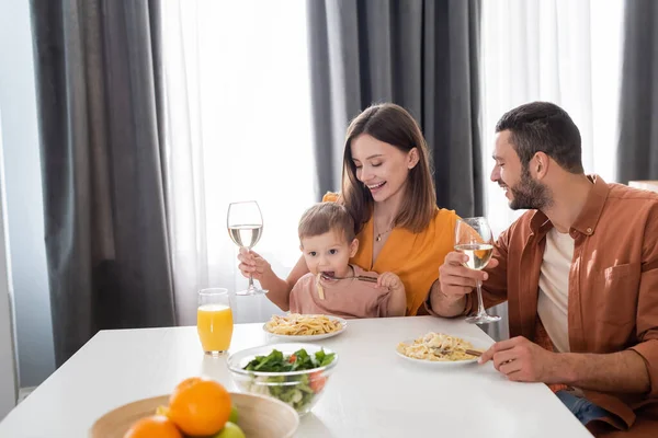 Mujer alimentando a su hijo y sosteniendo el vino cerca de marido y pasta en casa - foto de stock