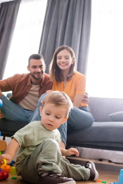 Toddler boy playing building blocks near blurred parents on couch in living room — Stock Photo