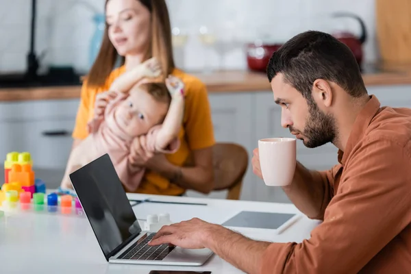 Freelancer holding cup and using laptop near family and building blocks at home — Stock Photo