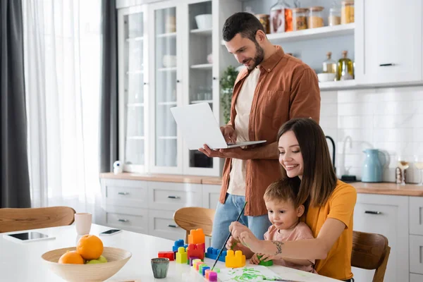 Junge Frau nähert sich Sohn und Ehemann mit Laptop in Küche — Stockfoto