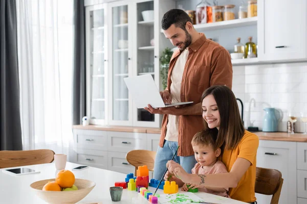 Mujer joven que se acerca al hijo y el marido utilizando el ordenador portátil en la cocina - foto de stock