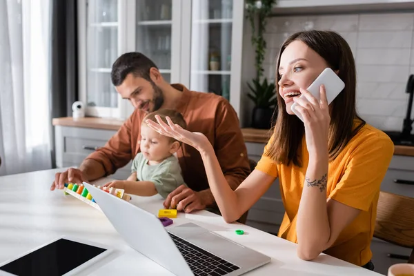 Jovem mulher falando no smartphone perto de gadgets e família em casa — Fotografia de Stock