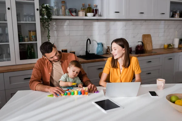 Man and kid playing game near mother working on laptop and fruits at home — Stock Photo