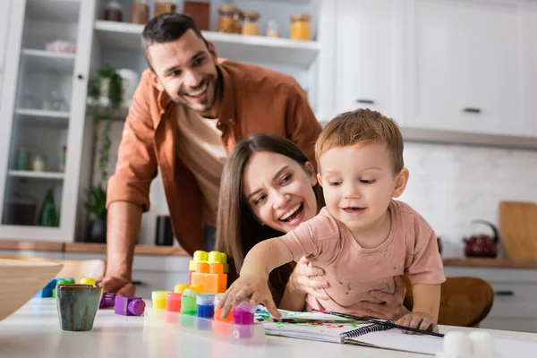 Bambino sorridente che prende vernice vicino sketchbook e genitori a casa — Foto stock