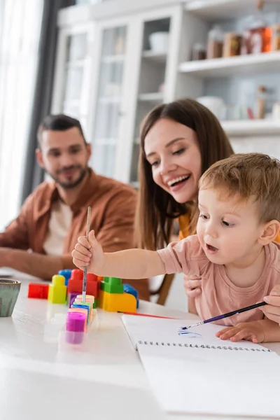 Ragazzo che tiene pennello vicino sketchbook e mamma a casa — Foto stock