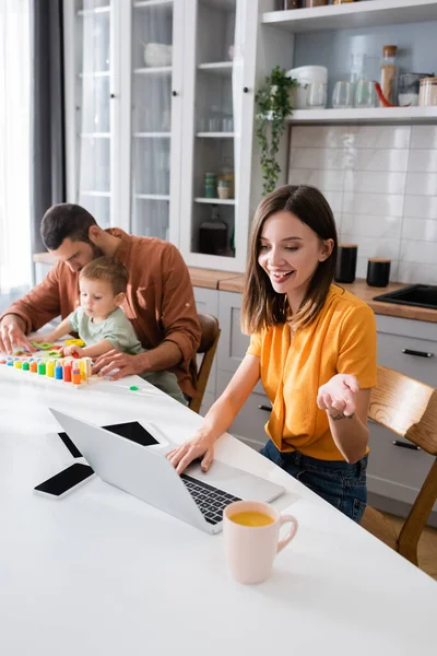 Freelancer having video call on laptop near coffee and family at home — Stock Photo