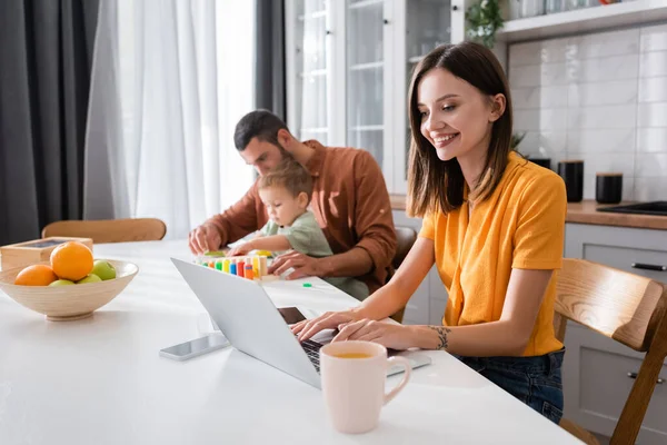 Un pigiste souriant utilisant un ordinateur portable près du café, des fruits et de la famille à la maison — Photo de stock