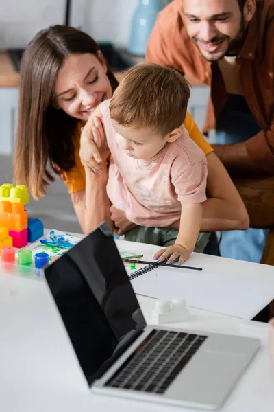 Portátil cerca de la familia sonriente, cuaderno de bocetos y bloques de construcción en casa - foto de stock