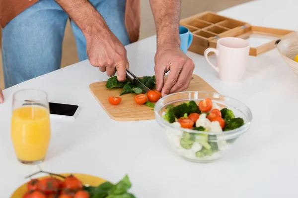 Vista recortada de hombre cortando verduras cerca de teléfono inteligente y jugo de naranja en casa - foto de stock