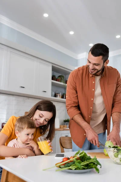 Hombre feliz cortando ensalada cerca de la esposa con un vaso de jugo de naranja e hijo en la cocina - foto de stock