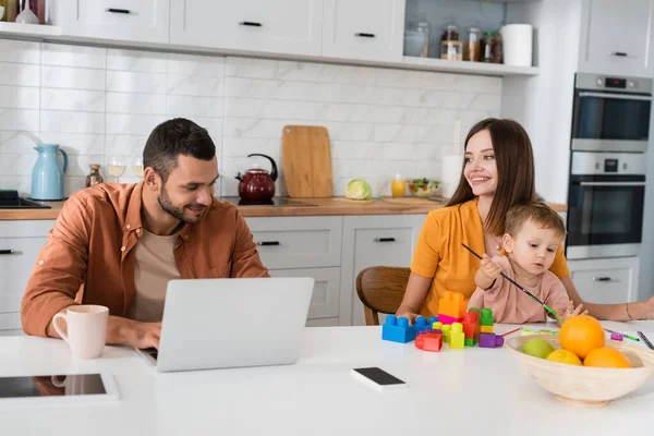 Frau und Sohn malen nahe Vater mit Laptop in Küche — Stockfoto