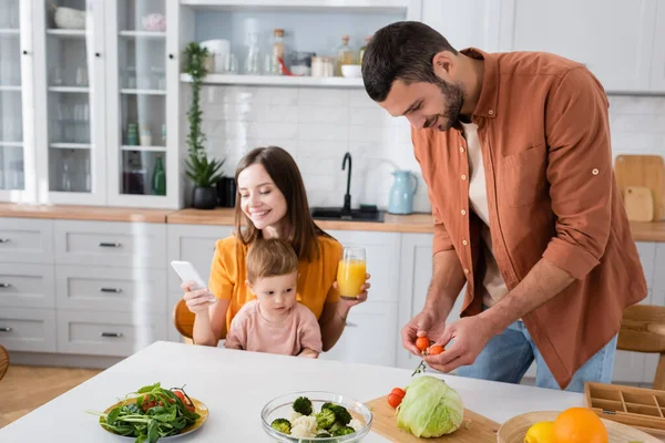 Young man holding cherry tomatoes near smiling wife using smartphone and son at home — Stock Photo